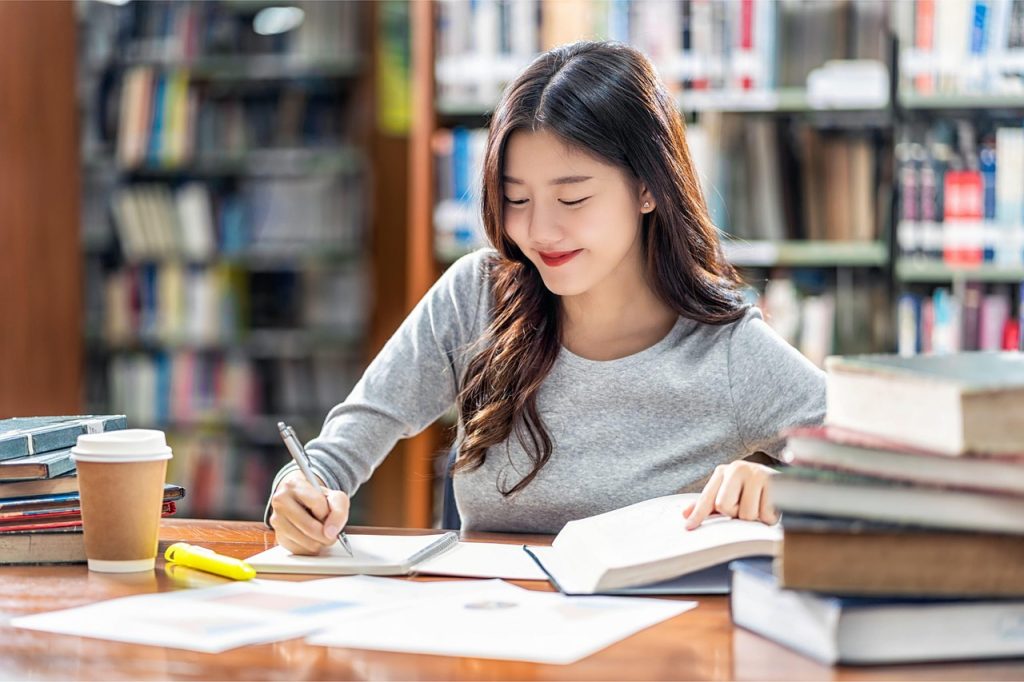 Academic Writing Help UK Student sitting in a library writing notes with open books on the desk - Academic Writing Help UK