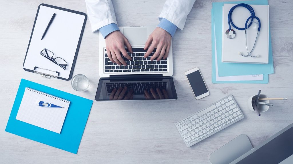 Nursing Assignment Help UK – nurse typing on laptop with stethoscope, notes and referencing tools